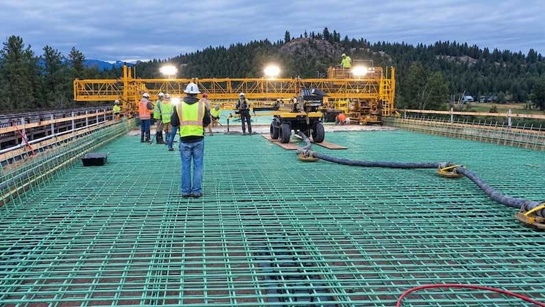 Crews place concrete for the deck of the new Sportsman’s Bridge over the Flathead River on Montana Highway 82 near Bigfork. Workers in high-visibility safety gear stand on a grid of reinforcing steel while a concrete placement machine operates behind them.