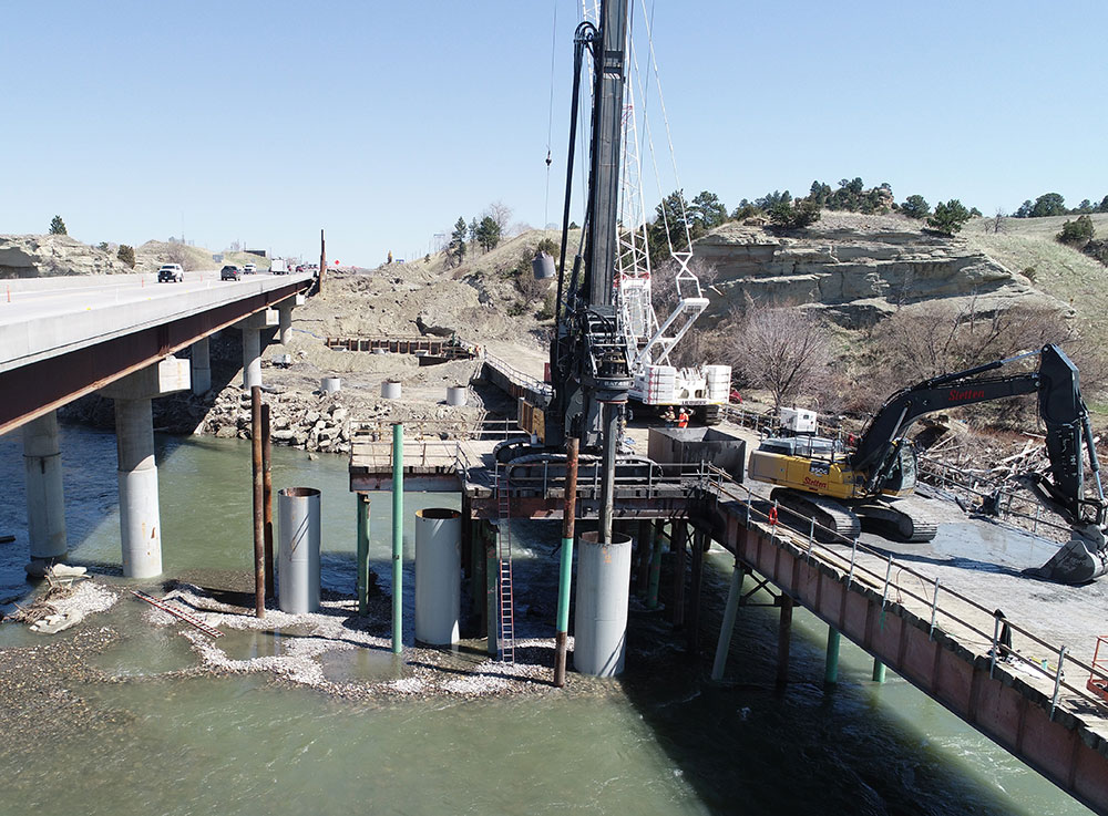 Crews and equipment drilling below the riverbed to build the new concrete foundations for the eastbound bridge.